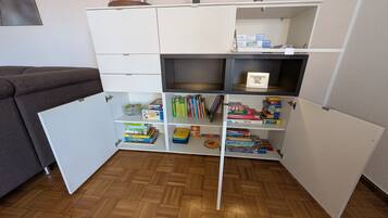 A room with a wooden herringbone floor, a white cabinet with open shelves containing books and board games, and a grey sofa.