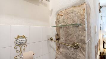 Un baño con pared de piedra, toallero, repisa de vidrio y dispensador de papel higiénico con un letrero decorativo.