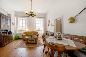 Une pièce avec un plancher en bois, un canapé, une table à manger avec des chaises, un lustre et une grande fenêtre.