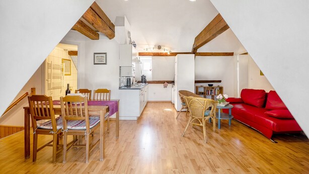Een zolderkamer met een puntdak, houten vloeren, een eettafel met stoelen, een rode bank en een keuken in de achtergrond.