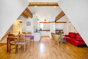 Habitación ático triangular con suelo de madera, una mesa de comedor con sillas, un sofá rojo y una zona de cocina al fondo.