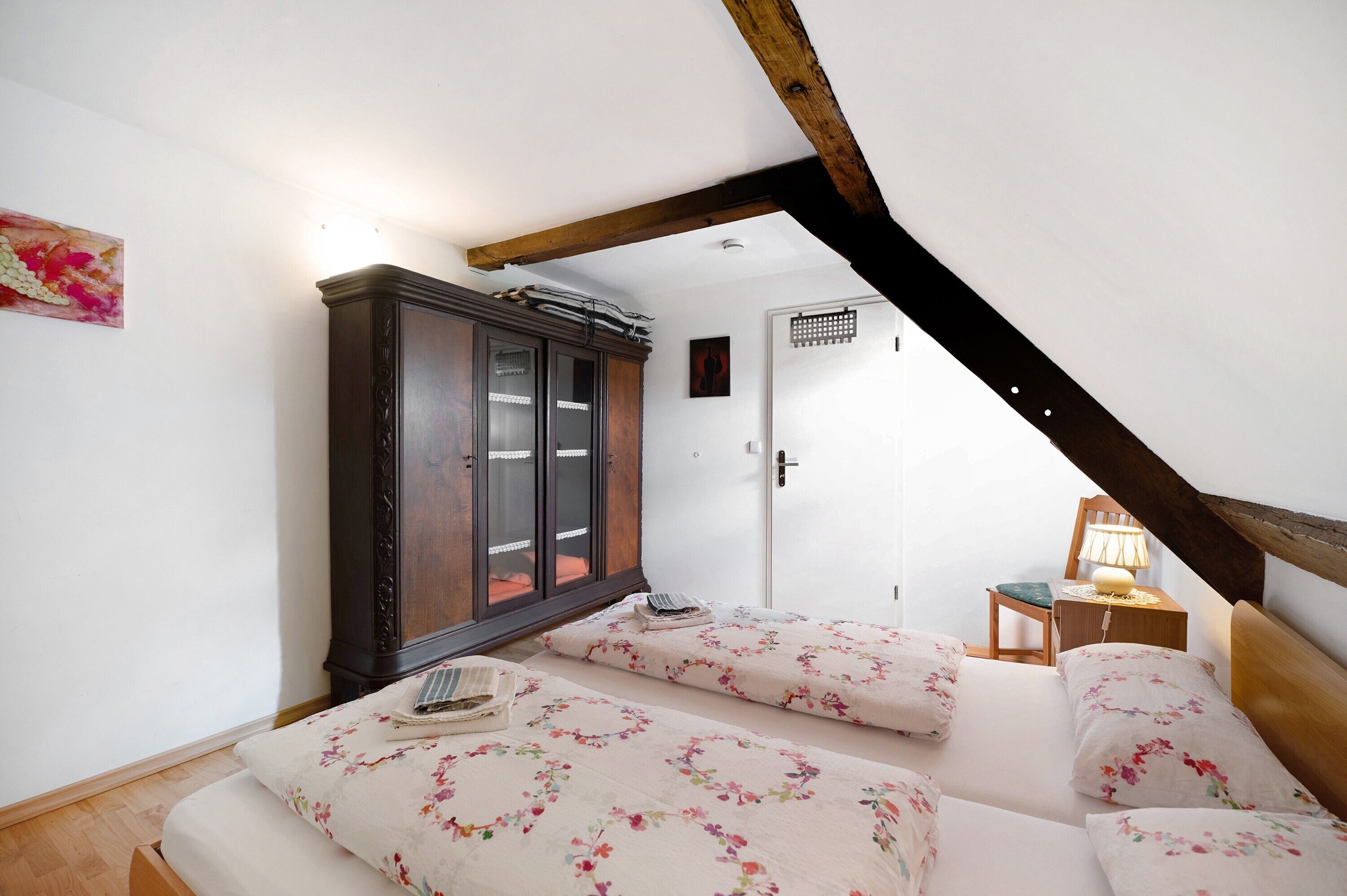 Un dormitorio con techo de madera, un armario, una cama con ropa de cama floral y una mesita de noche con una lámpara.
