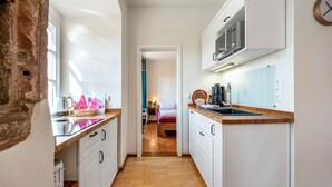 Eine schmale Küche mit weißen Schränken, einer Holz Arbeitsplatte und einer Mikrowelle. Ein Holzboden führt zu einem Schlafzimmer mit einem Bett und einem Nachttisch.