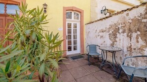 Um edifício amarelo com uma porta branca, uma pequena área de estar ao ar livre com duas cadeiras e uma mesa, e um vaso com planta em primeiro plano.