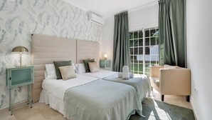 A hotel room with two beds, a nightstand with a lamp, a small cabinet, and a view of the outdoors through a window with curtains. - VILLA FALDO VACATION MARBELLA (Nueva Andalucía)