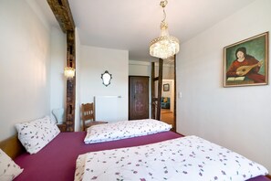 A bedroom with two beds, a chandelier, a painting, and a view into another room.