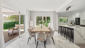 A modern kitchen with a marble countertop, a dining area with a wooden table and upholstered chairs, and a view of a lush garden through large windows.