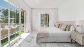 A bedroom with a large bed, a nightstand, a lamp, and a view of greenery through the windows.