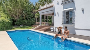 A swimming pool with a clear blue water, surrounded by a white building with a balcony and steps leading to a covered patio area. - VILLA NADAL VACATION MARBELLA (Elviria)