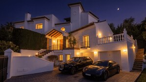 Uma casa de dois andares com entrada por uma escadaria, um caminho de acesso e dois carros estacionados.