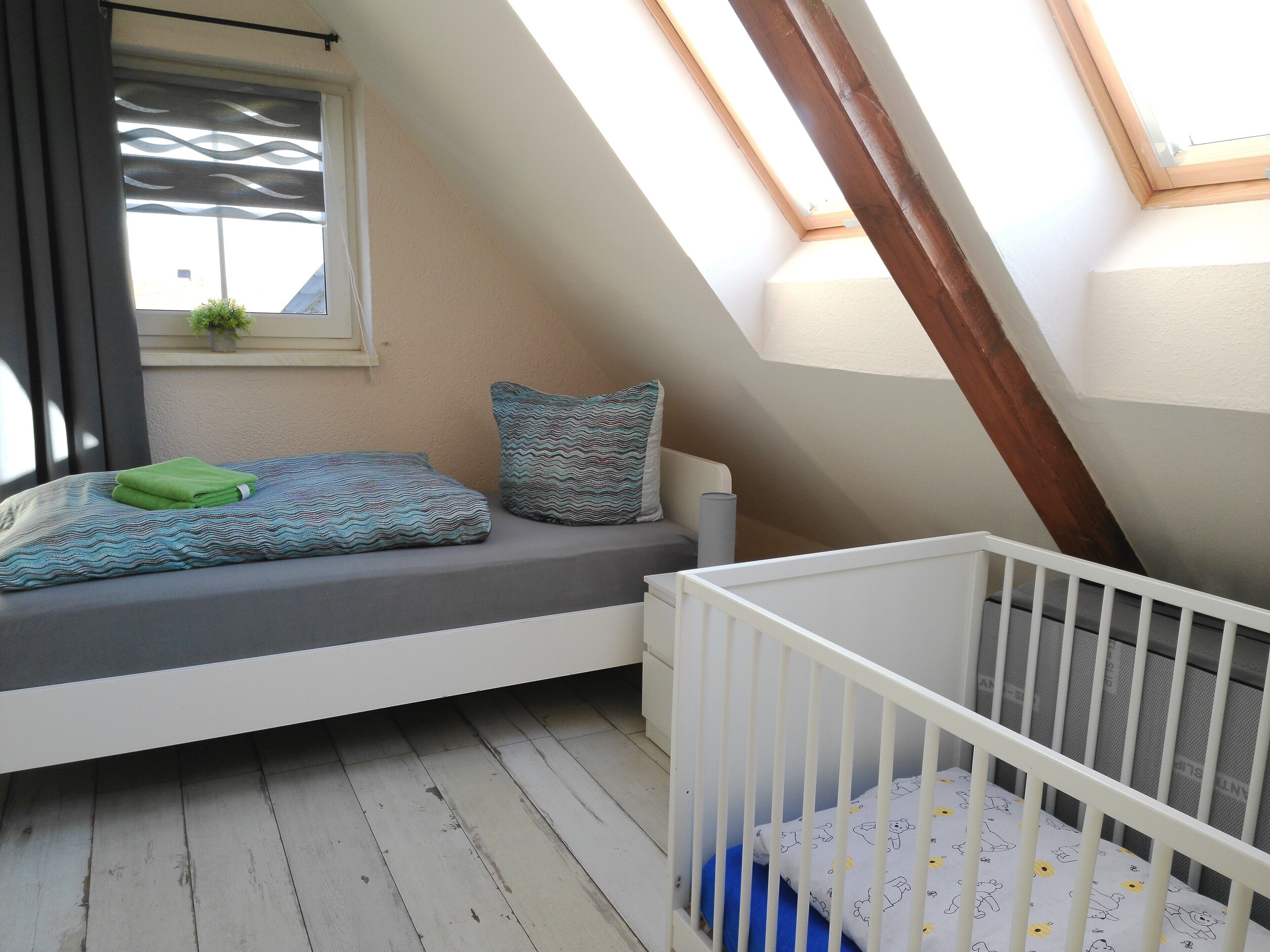 A room with a sloped ceiling, a window with a curtain, a small bench with pillows, and a crib.