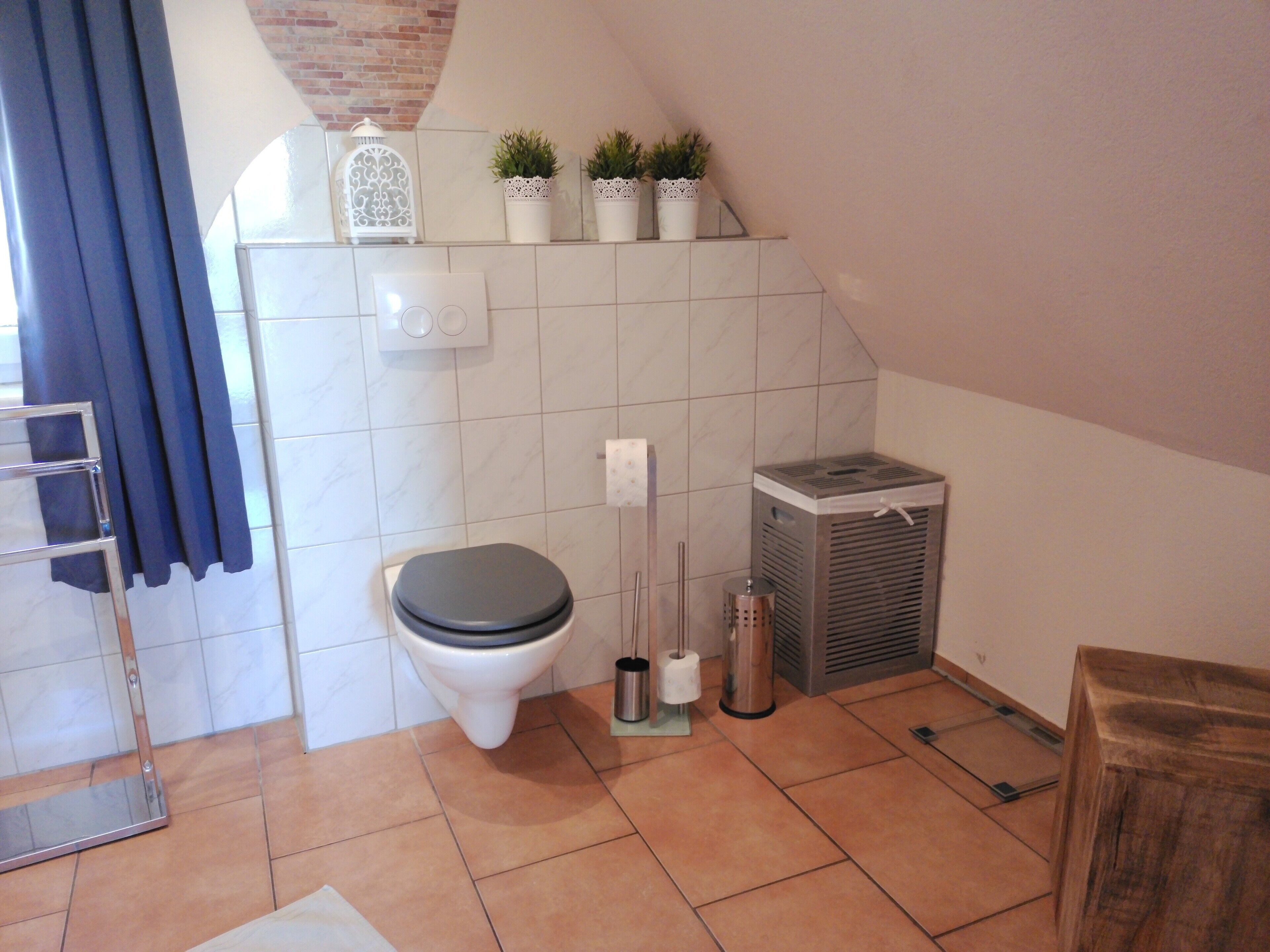 A bathroom with a toilet, a hand dryer, a trash can, a shelf with plants, and a shower area.