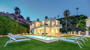 A modern two-story house with a swimming pool, surrounded by a well-maintained lawn and palm trees. - VILLA MONTE HALCONES VACATION MARBELLA (BENAHAVIS)