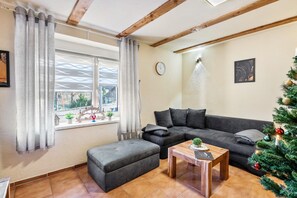 A living room with a grey sofa, a wooden coffee table, a grey ottoman, a window with curtains, a wall clock, and a Christmas tree.