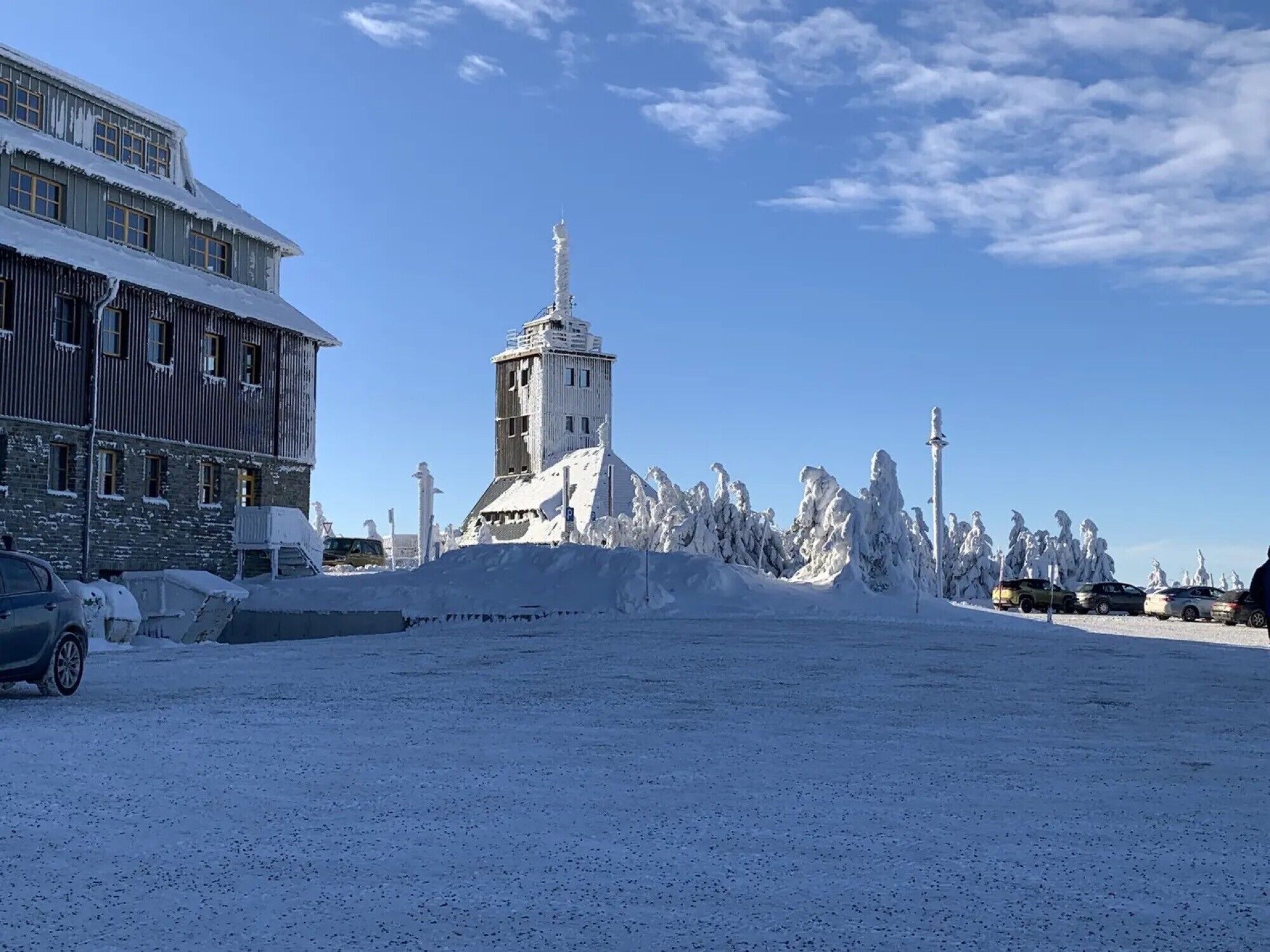 A snow-covered building with a prominent tower, parked cars, and a clear sky.