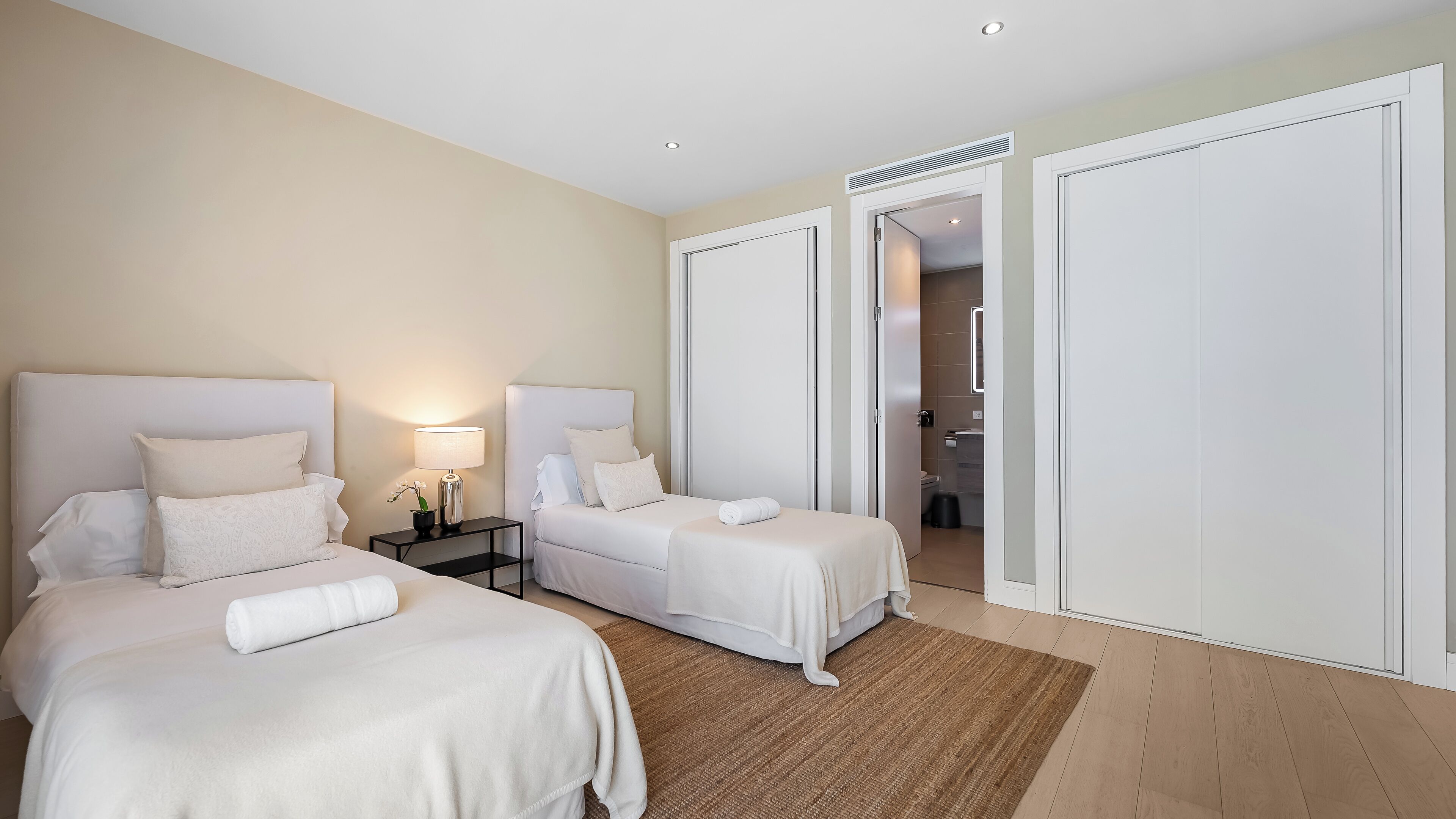 A hotel room with two beds, a nightstand with a lamp, a bathroom visible through an open door, and a wooden floor.