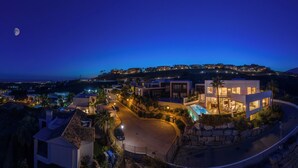 Eine moderne Villa mit Pool, umgeben von einer Steinmauer und Palmen, mit Blick auf eine Küstenstadt in der Nacht.