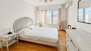 A bedroom with a bed, bedside table, dresser, window with curtains, and a view of buildings. - Comfortable and modern two bedroom (Cannes)