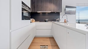 Een moderne keuken met witte kasten, een accentmuur van donker hout en uitzicht op zee.