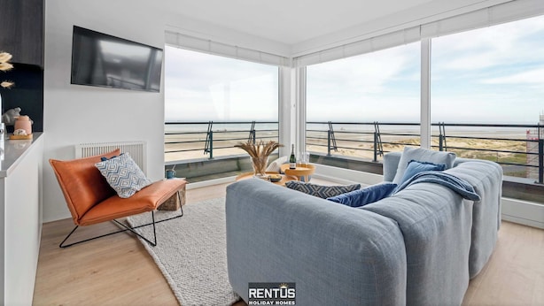 Een moderne woonkamer met een grote grijze bank, een bruine leren fauteuil en een flatscreen televisie aan de muur bevestigd. De kamer heeft grote ramen met uitzicht op het strand en de zee.