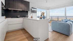 Een moderne keuken met een centraal eiland, een zithoek met een bank en een groot raam met uitzicht op zee.