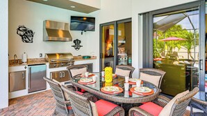 An outdoor kitchen with a grill, a TV, and wicker chairs around a glass table.