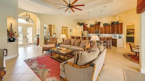 A spacious living room with a large sofa, a coffee table, and a red rug. There is a kitchen area with wooden cabinets and a dining area with chairs.