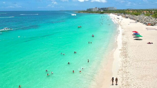 Une plage avec une eau turquoise et limpide, des gens qui se baignent et des parasols sur le sable.