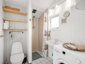 Ein kleines Badezimmer mit Toilette, Waschbecken und Waschmaschine. Über der Toilette befinden sich Ablagen für Pflegeprodukte, und im Hintergrund ist eine Dusche zu sehen.