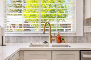 A kitchen with a white countertop, a gold faucet, a soap dispenser, and a window with blinds. - Upscale 5BR/3BA w/ Jacuzzi (Oak Park)