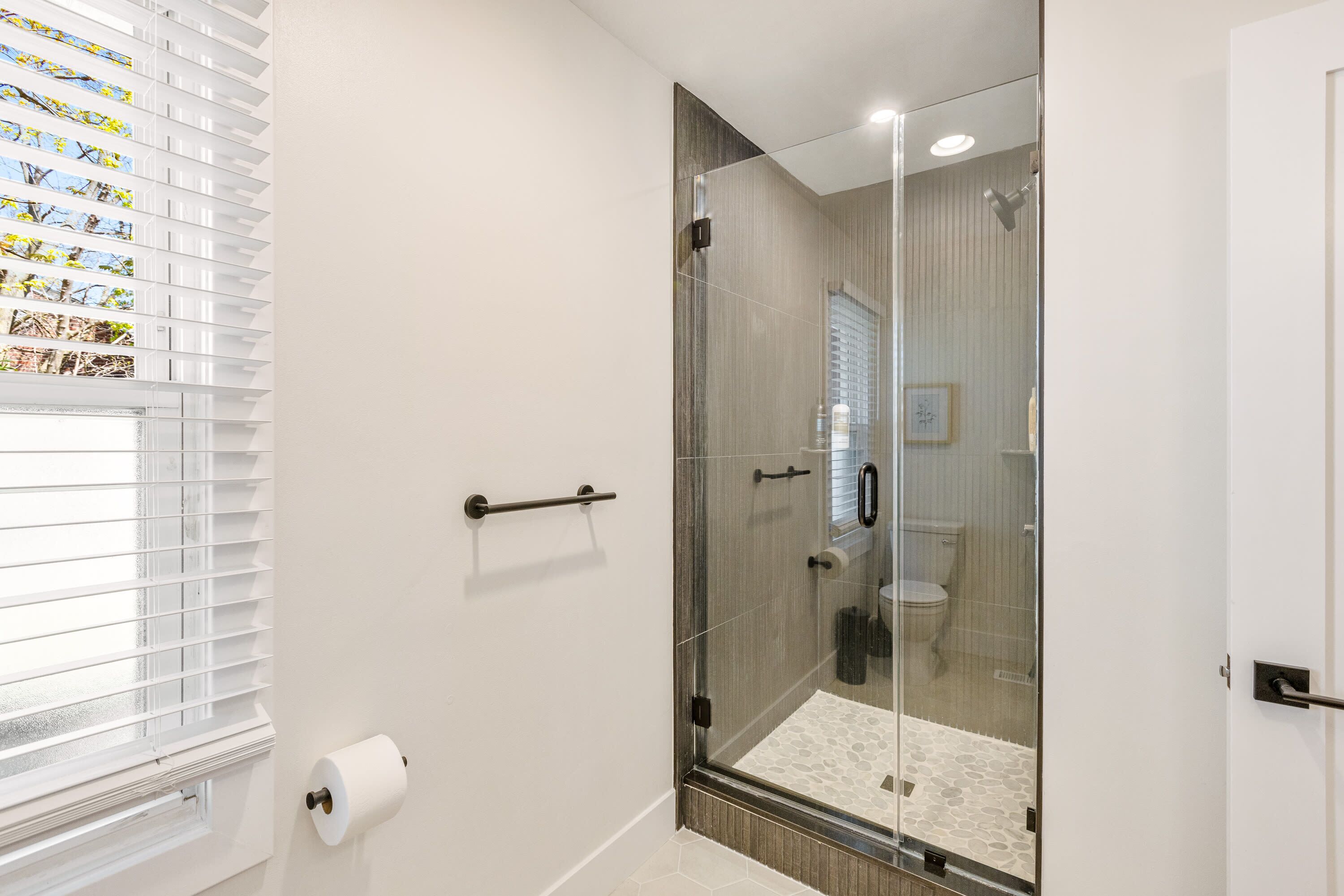 A bathroom with a glass shower enclosure, a toilet, and a window with blinds.