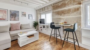 Ett modernt vardagsrum med en soffa, ett soffbord och två barstolar. Det finns en tv på en vit hylla, ett fönster med gardiner och en tegelvägg.