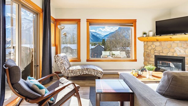 A living room with a stone fireplace, two large windows with mountain views, a flat-screen TV, and a cozy seating area.