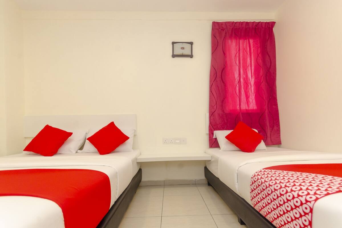 Habitación de hotel con dos camas, cada una con ropa de cama y almohadas rojas, una mesita de noche pequeña y una ventana con cortinas rosadas.