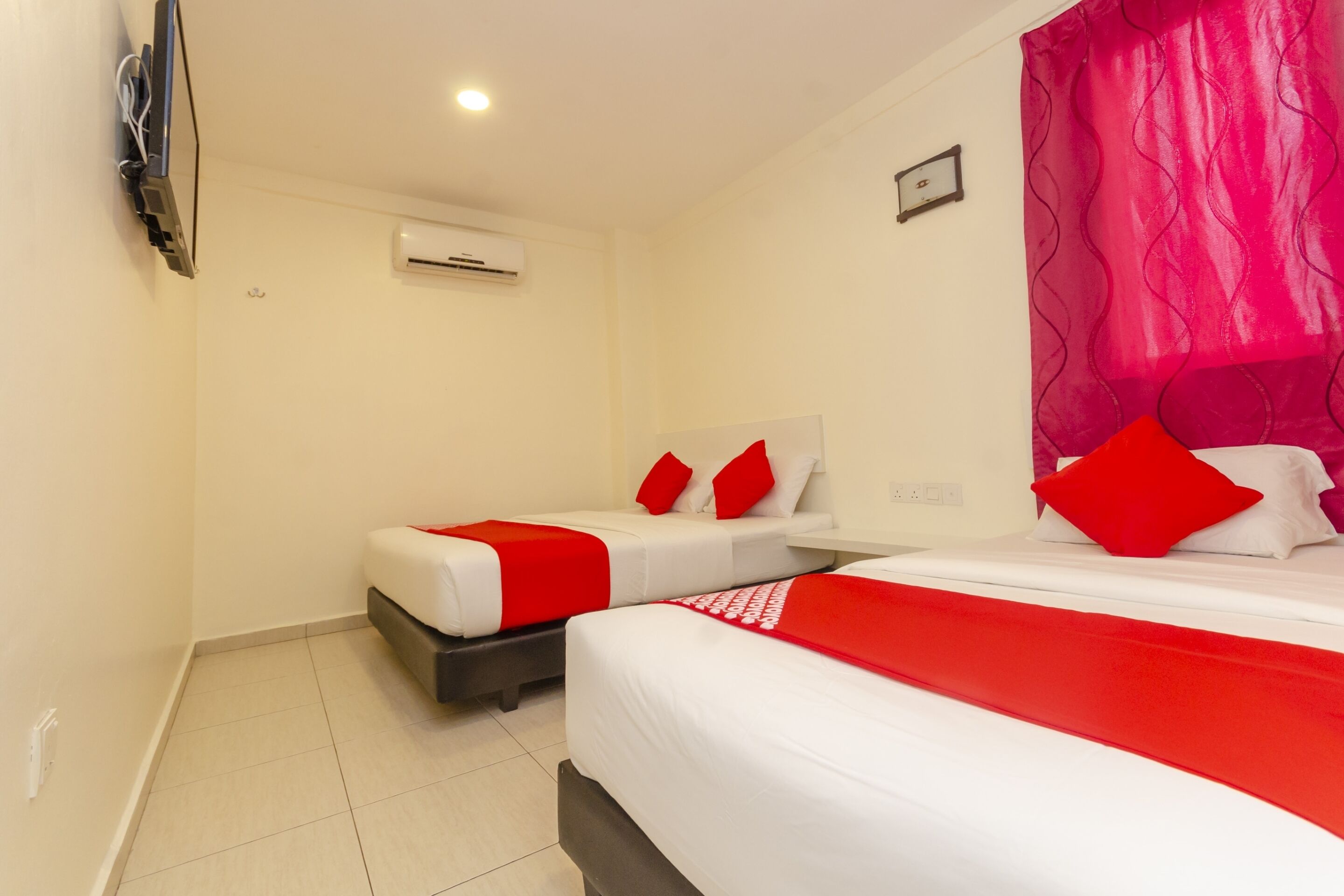 A hotel room with two beds, each with red and white bedding, a TV mounted on the wall, and an air conditioning unit.