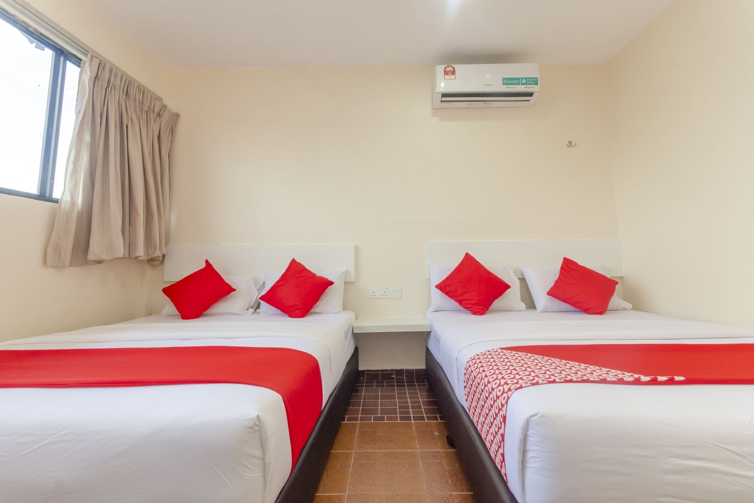 Habitación de hotel con dos camas, cada una con ropa blanca y almohadas rojas. Se observa una unidad de aire acondicionado en la pared, una ventana con cortinas color beige y piso de baldosas.