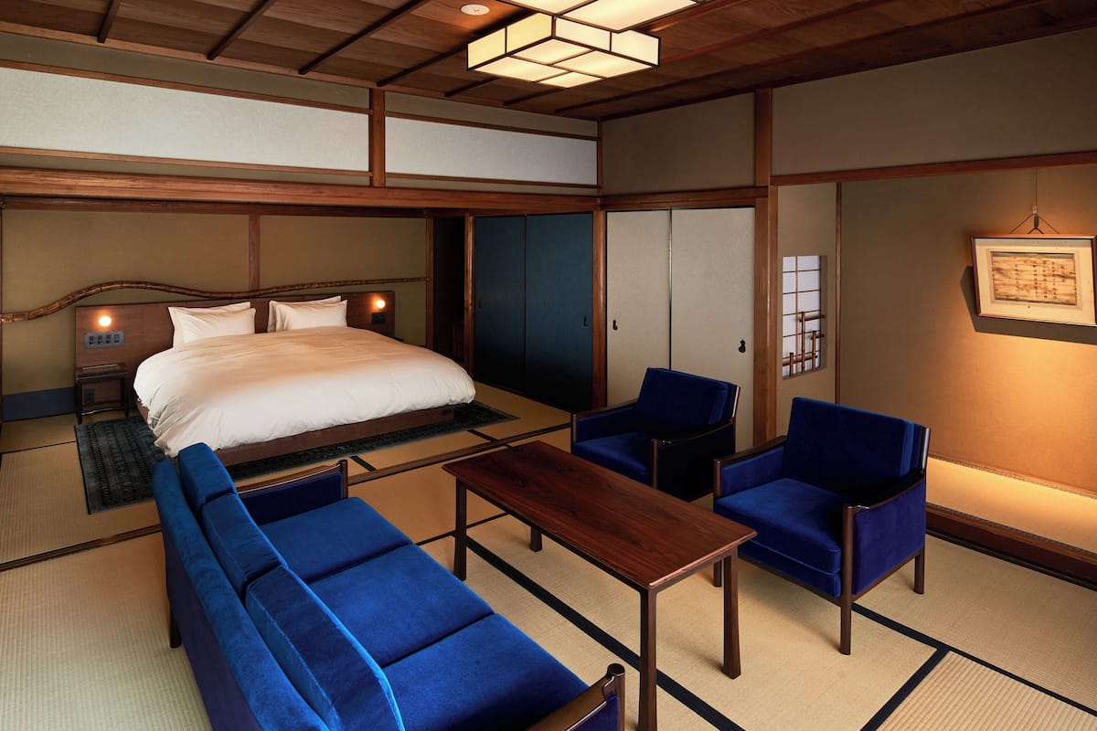 A traditional Japanese-style room with tatami flooring, a bed with white bedding, two blue upholstered chairs, a wooden table, and a ceiling light fixture.