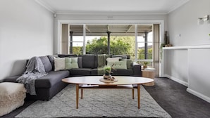 Un salon moderne avec un canapé d’angle gris, une table basse ronde et une vue sur la verdure grâce à de grandes fenêtres.