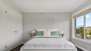 Une chambre avec un lit blanc, deux éléments de décoration murale assortis, une fenêtre avec vue et une armoire blanche.
