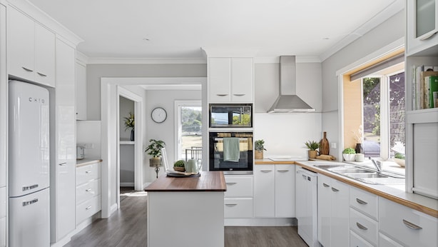 Une cuisine moderne avec des meubles blancs, un îlot central et une vue sur l’extérieur grâce à une grande fenêtre.