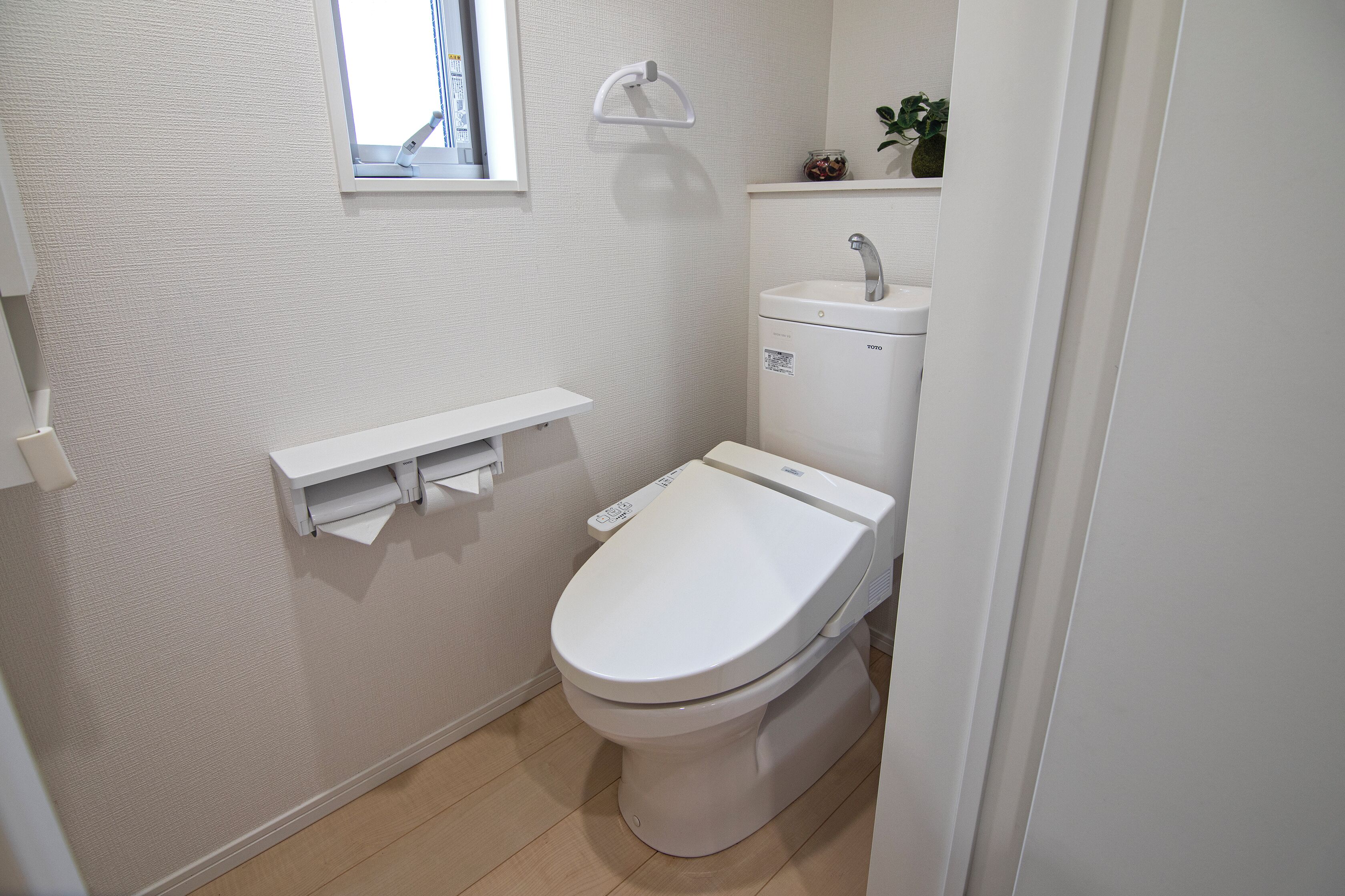 A small, clean bathroom with a toilet, a shelf with toilet paper, a window, and a sink with a faucet.