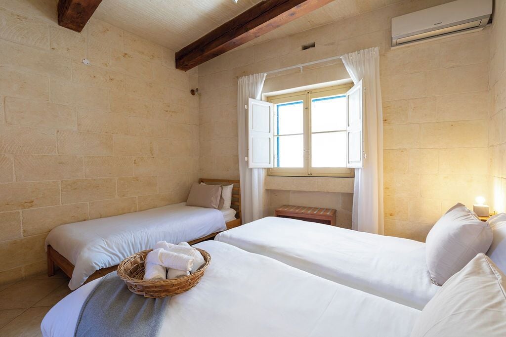 A room with two single beds, a window with curtains, a wall-mounted air conditioner, and a small bedside table with a lamp.