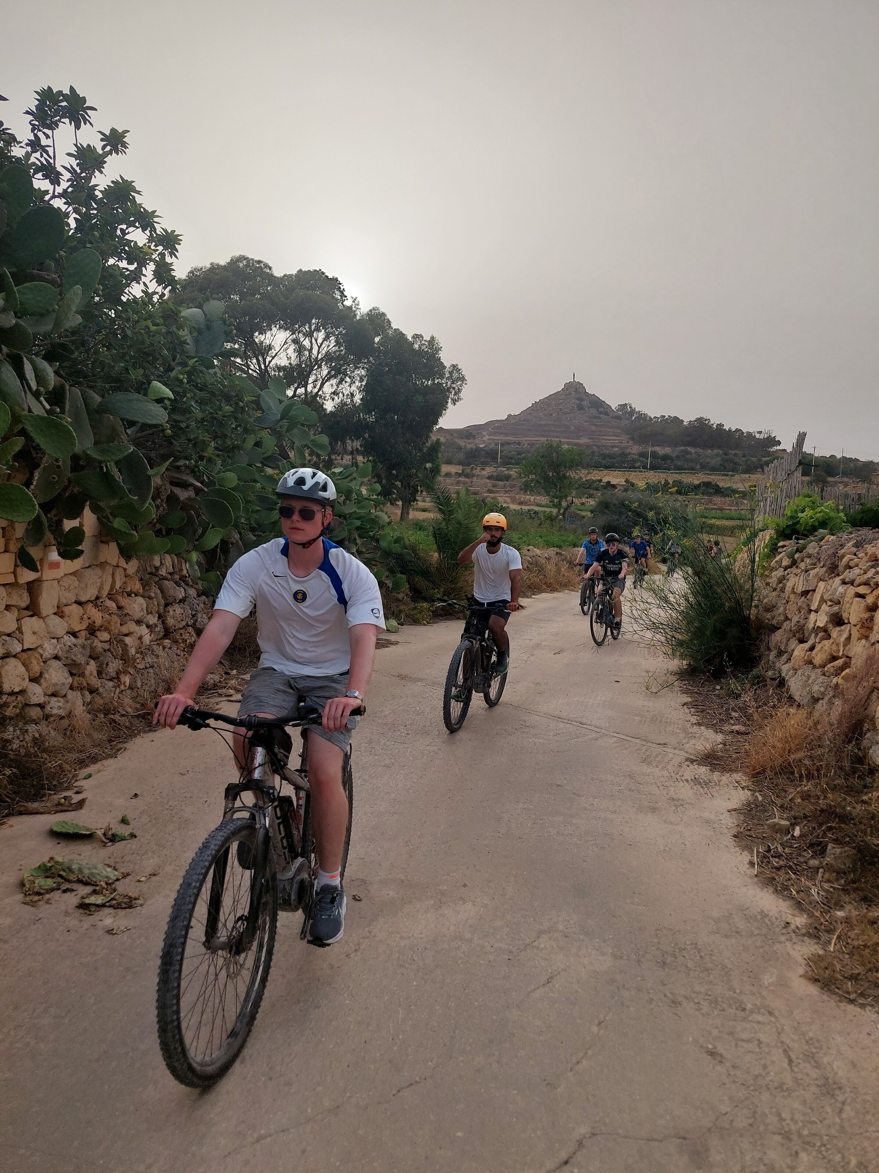 A group of cyclists on a paved path with stone walls and vegetation on either side.