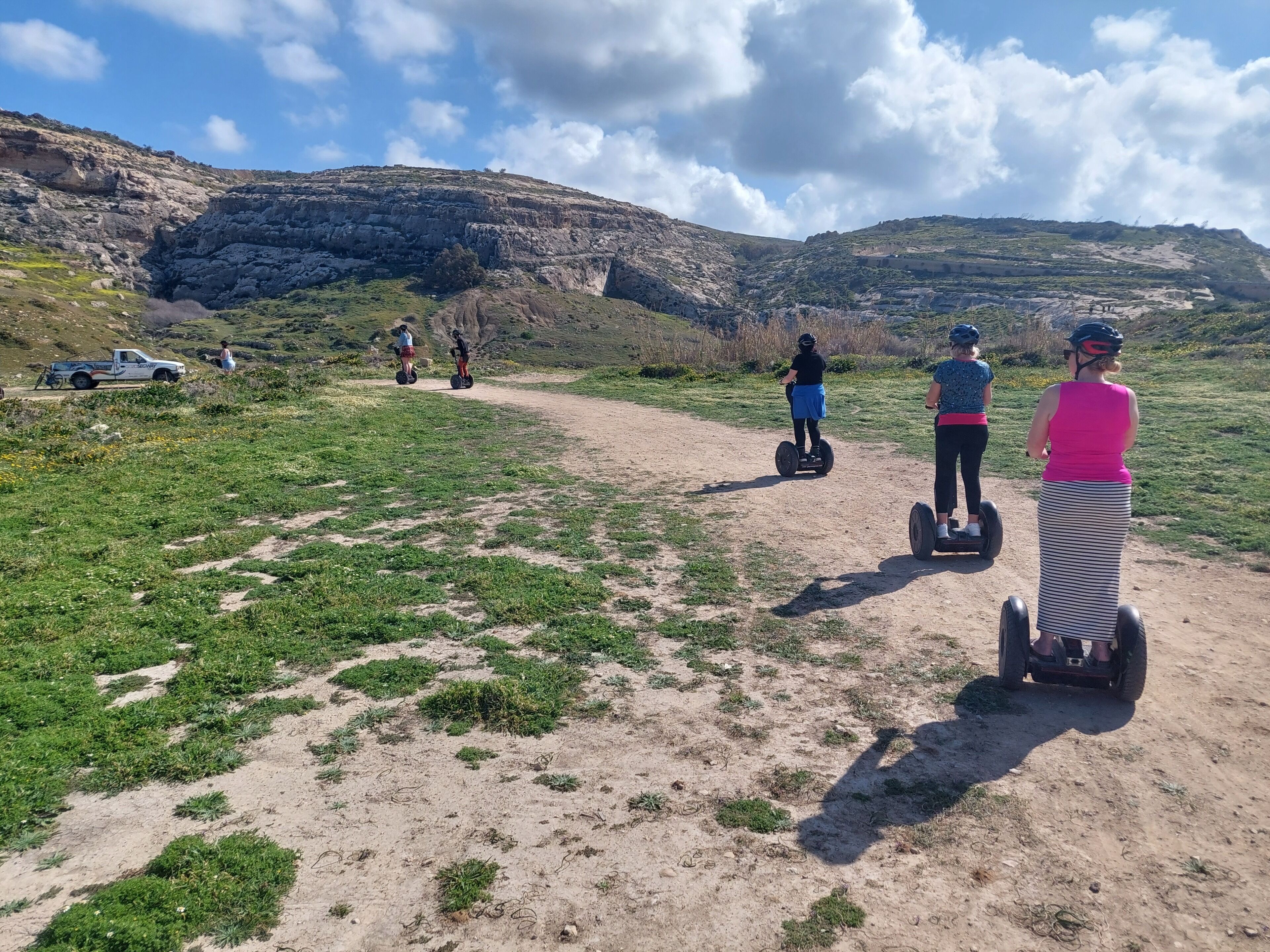 En grusväg med folk som åker p å segways, en vit pickup och en bergig sluttning i bakgrunden.
