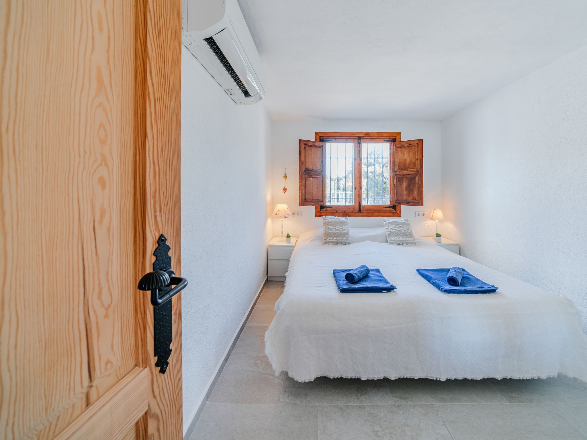 A bedroom with a bed, a window with wooden shutters, a bedside lamp, and a wall-mounted air conditioner.