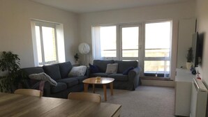 A living room with a sofa, a coffee table, and a dining table. There are windows with blinds, a plant, and a TV on the wall. - Central Spacious 3 Bedroom Apartment With Parking (Dublin)