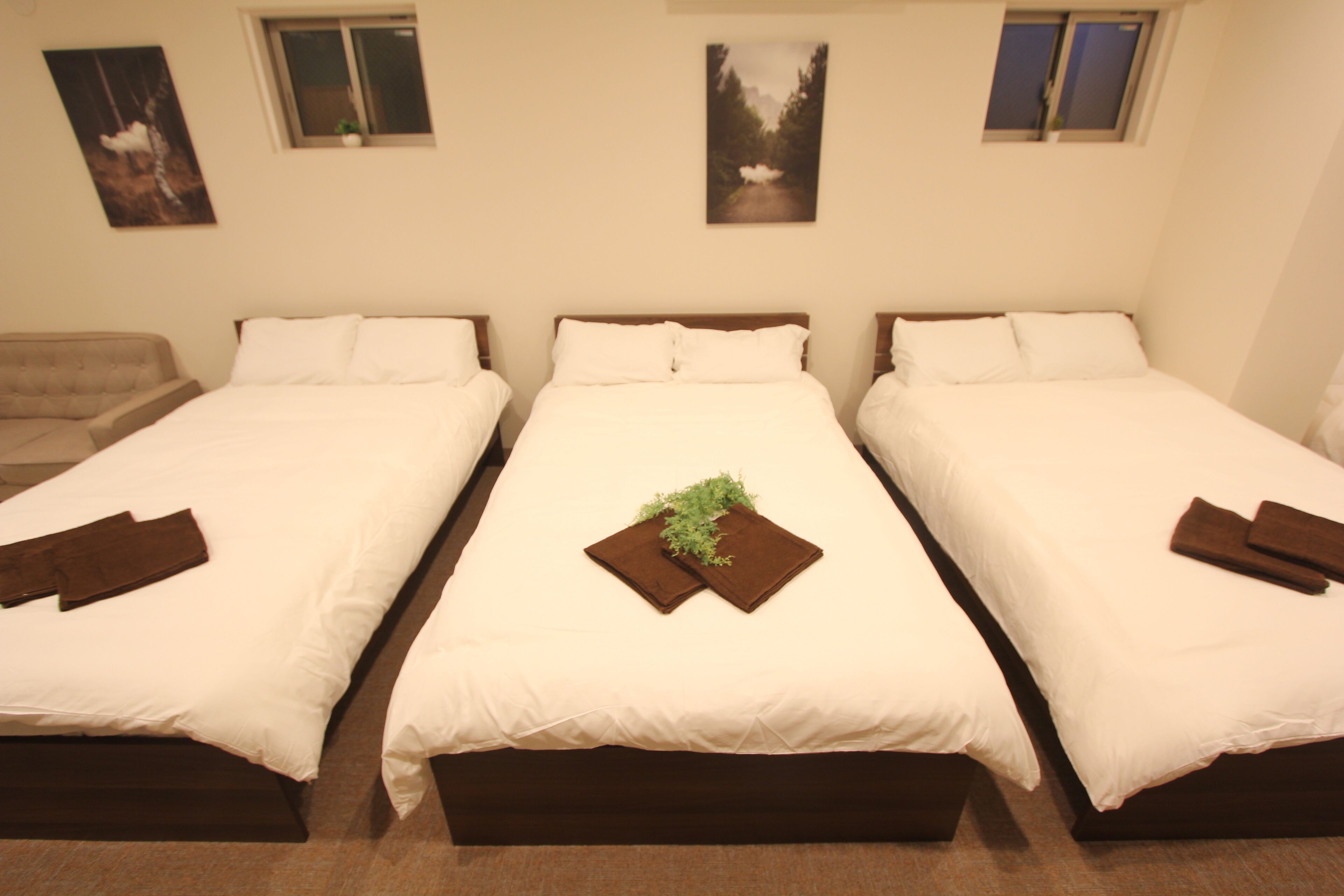 A hotel room with three single beds, each with white bedding and brown headboards. There are two windows on the wall, and artwork is displayed above the beds.