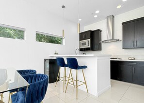A modern kitchen with a central island, bar stools, and a dining area with a glass table. - Stunning Modern Oasis - Heated Pool & Spa · Mountain Views · 5min to Downtown! (Palm Springs)