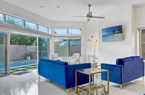 A modern living room with a glass coffee table, blue sofas, and a ceiling fan. - Stunning Modern Oasis - Heated Pool & Spa · Mountain Views · 5min to Downtown! (Palm Springs)