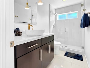 A modern bathroom with a white sink, dark cabinets, a glass shower enclosure, and a visible window. - Stunning Modern Oasis - Heated Pool & Spa · Mountain Views · 5min to Downtown! (Palm Springs)